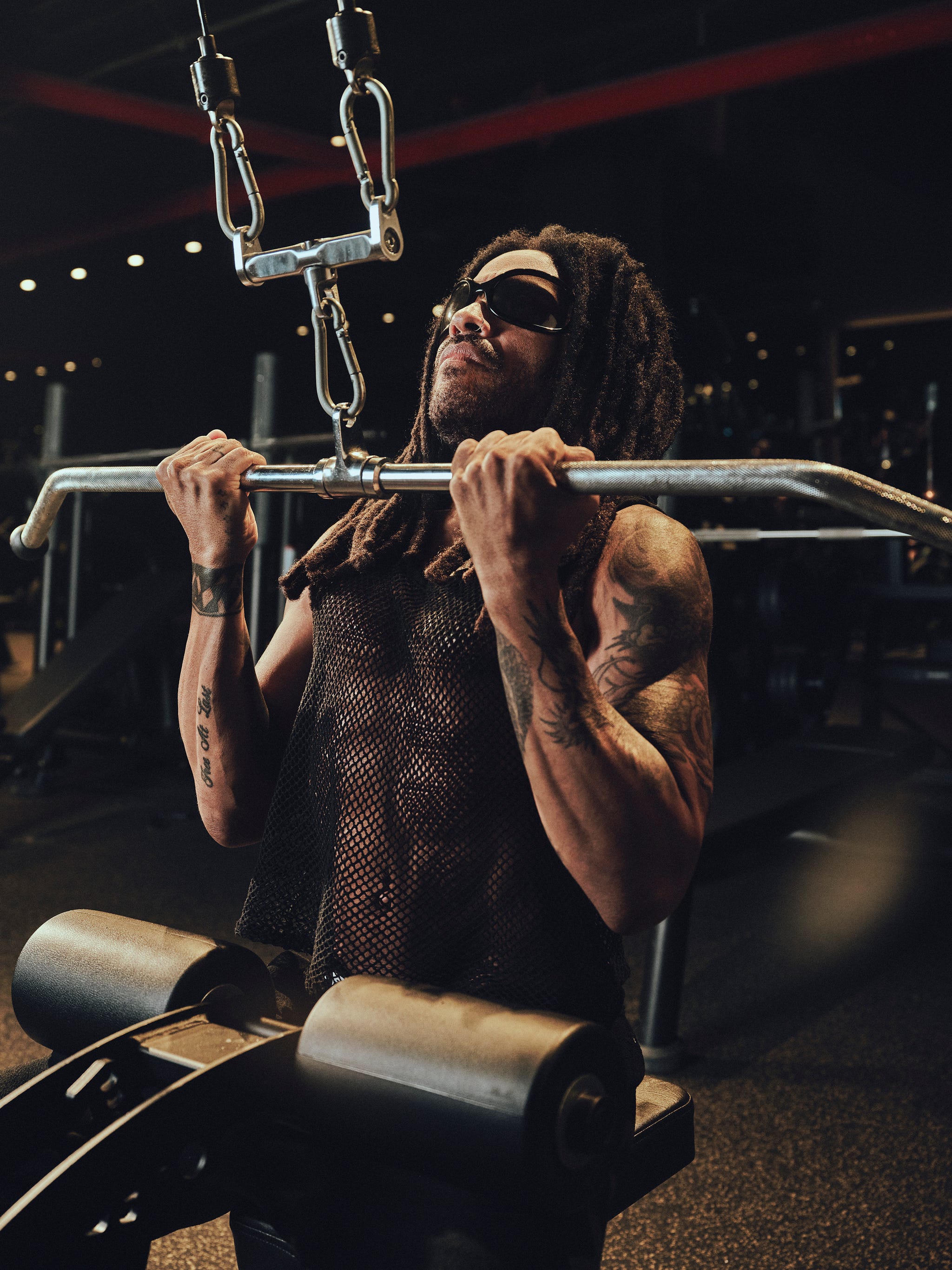 A person with dreadlocks performs a weightlifting exercise in a gym, wearing sunglasses and a mesh top.