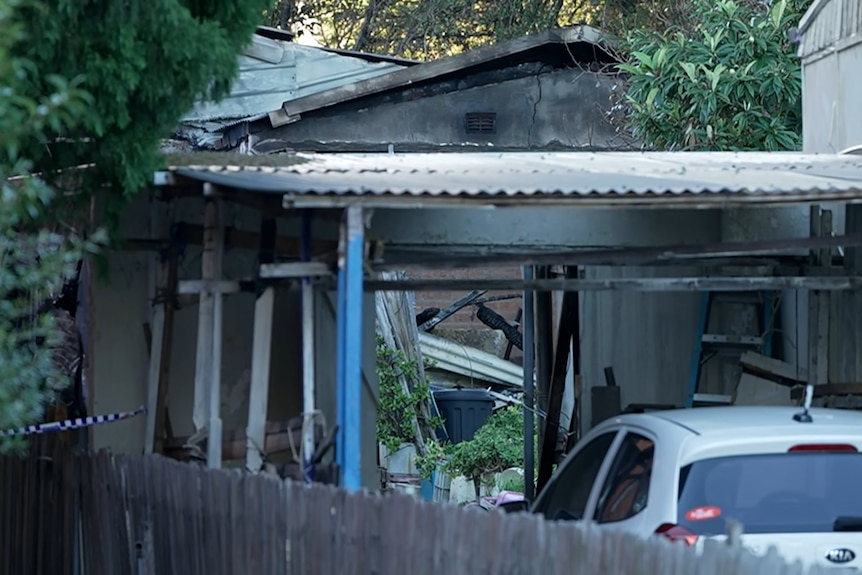 A white car parked in a driveway in front of a severly damaged granny flat which is mostly rubble.