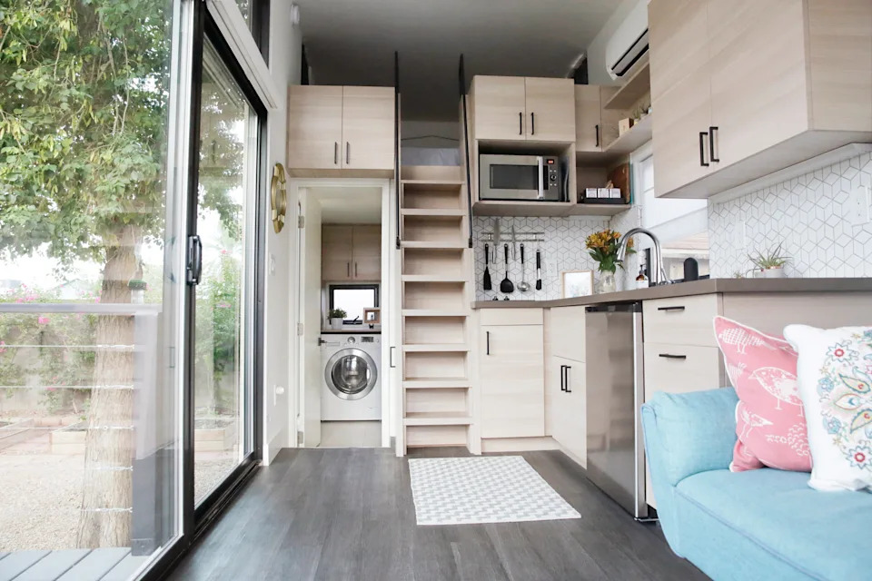 Modern tiny house interior with loft, compact kitchen, washing machine, and natural light through large glass doors. Cozy and efficient design