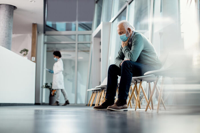 An older man wearing a face mask sits alone in a waiting room, reflecting on uncomfortable truths about the medical industry.