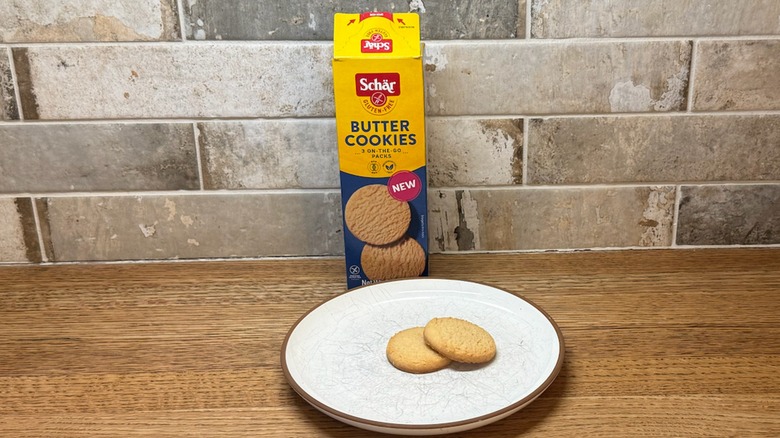 Two Schär Butter Cookies on a white plate with the whole box in the back leaning on a brick backsplash