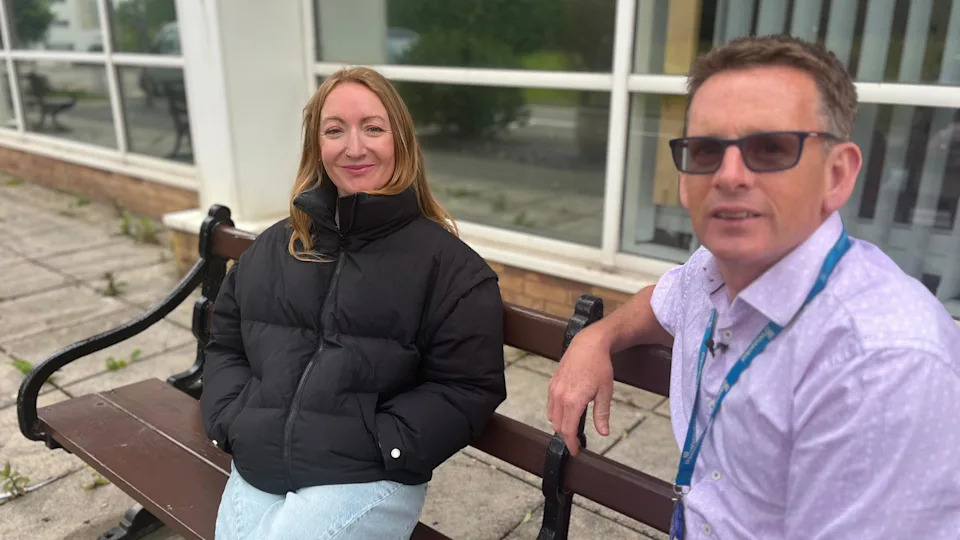 Lowri and Dr Healy sitting on a bench. Dr Healy is wearing a purple shirt with a lanyard, he has short dark hair and wears glasses. Lowri is wearing a black puffer jacket and jeans.