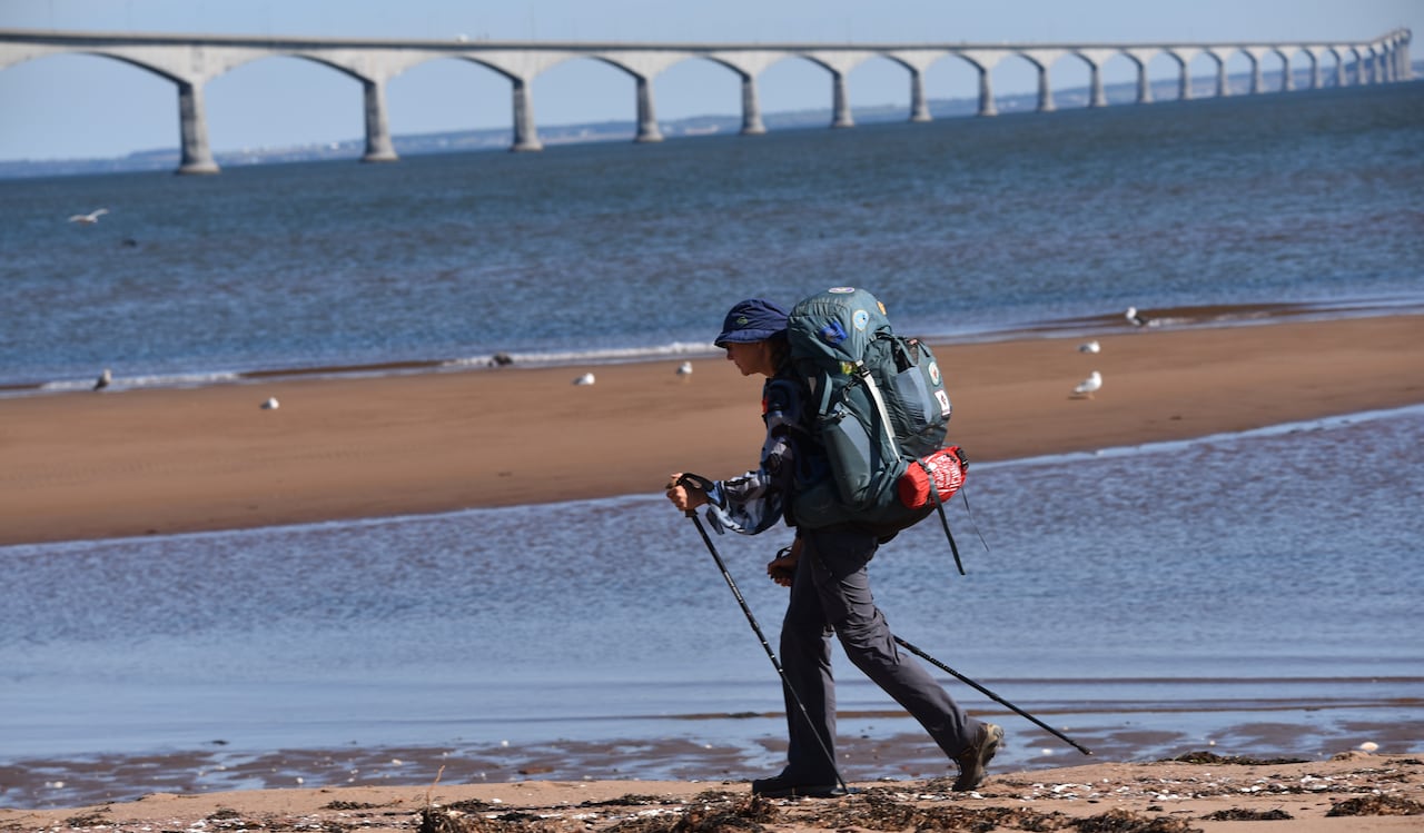 A woman wearing a large backpack walks on a sandy beach while a long bridge stretches in the background.