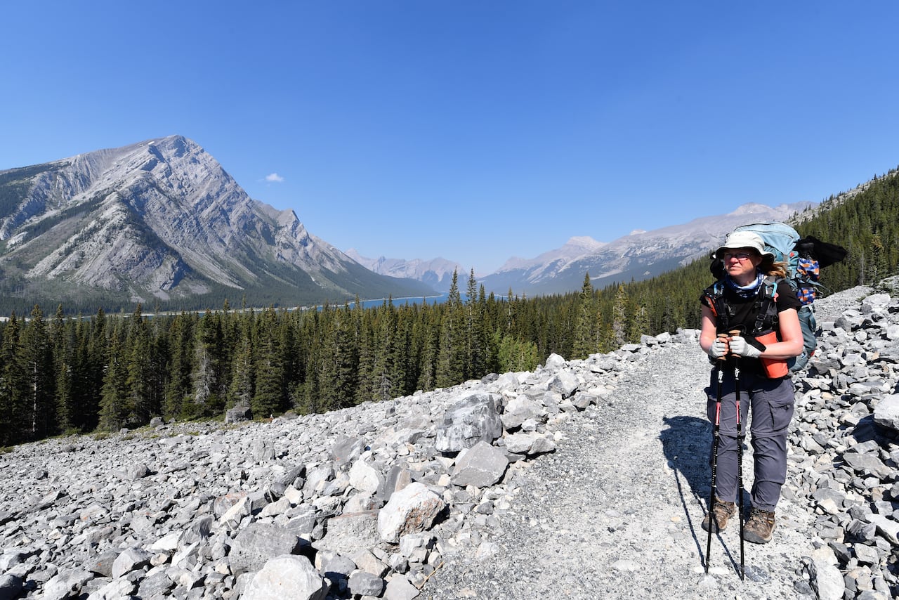 A woman stands on a rocky trail while looking at a view of mountains and a lake.