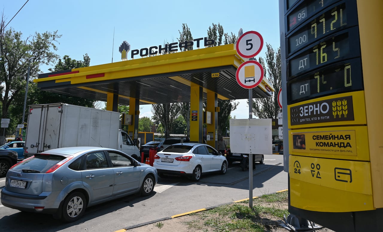 Vehicles are seen lining up in front of a gas station with cyrillic on it.