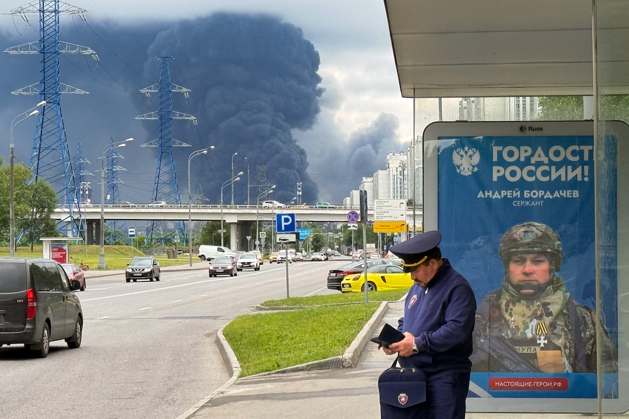 A man in a work uniform stands in front of a bus shelter while black smoke fills the sky above.