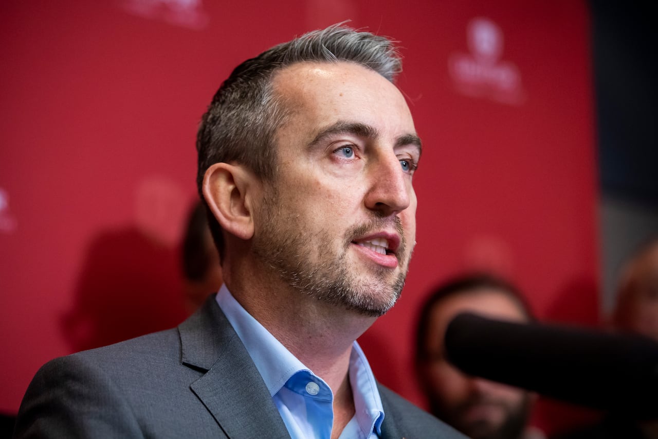 A man with a thin beard and wearing a grey suit jacket speaks in front of a red banner.