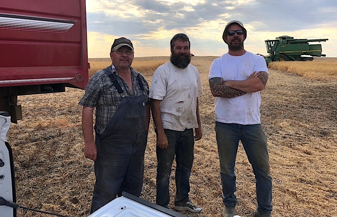 Three farms stand together in front of a field during harvest.