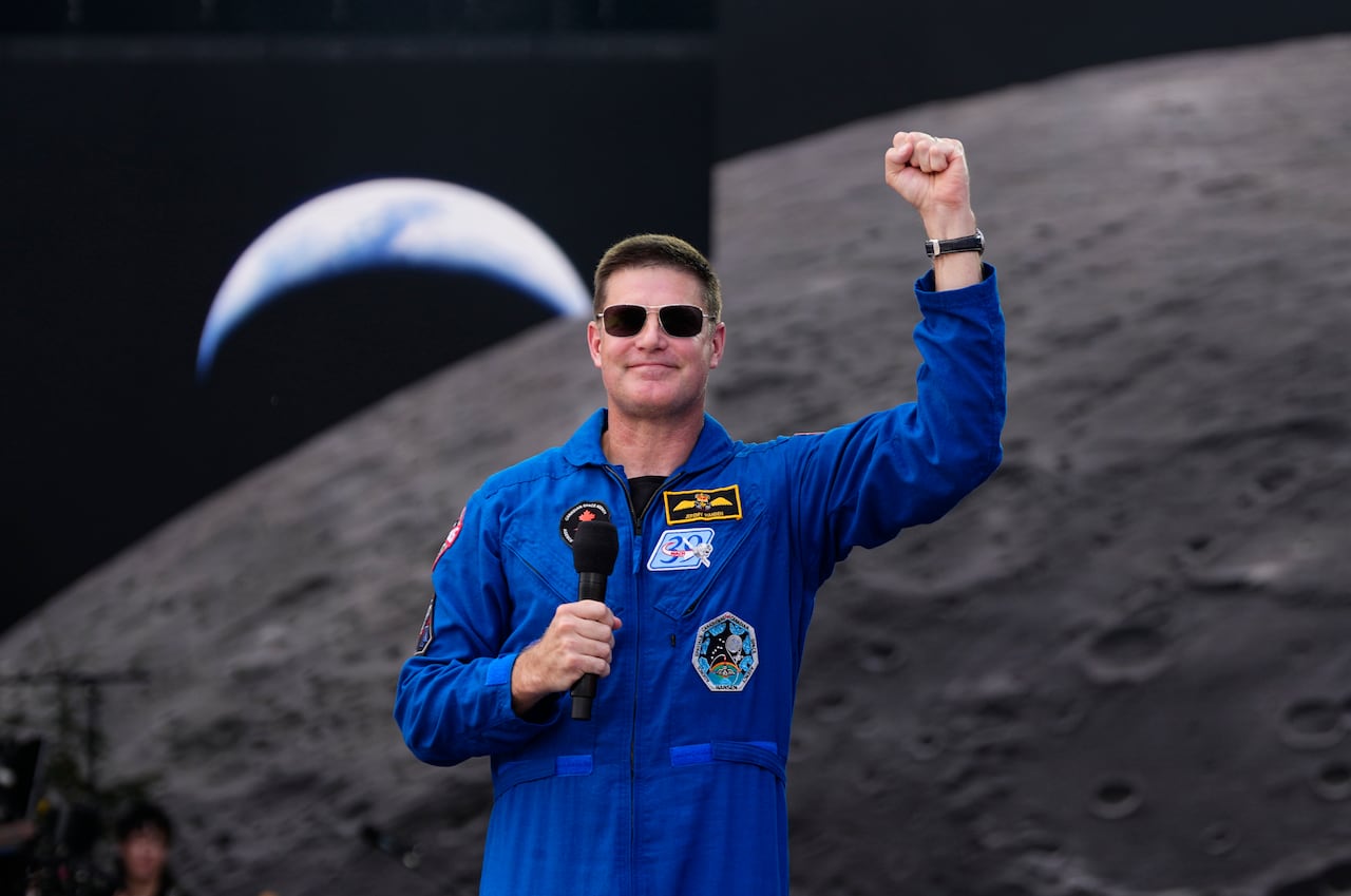 Man in sunglasses and a blue utility suit speaks with an arm raised and a silhouette of the moon behind him