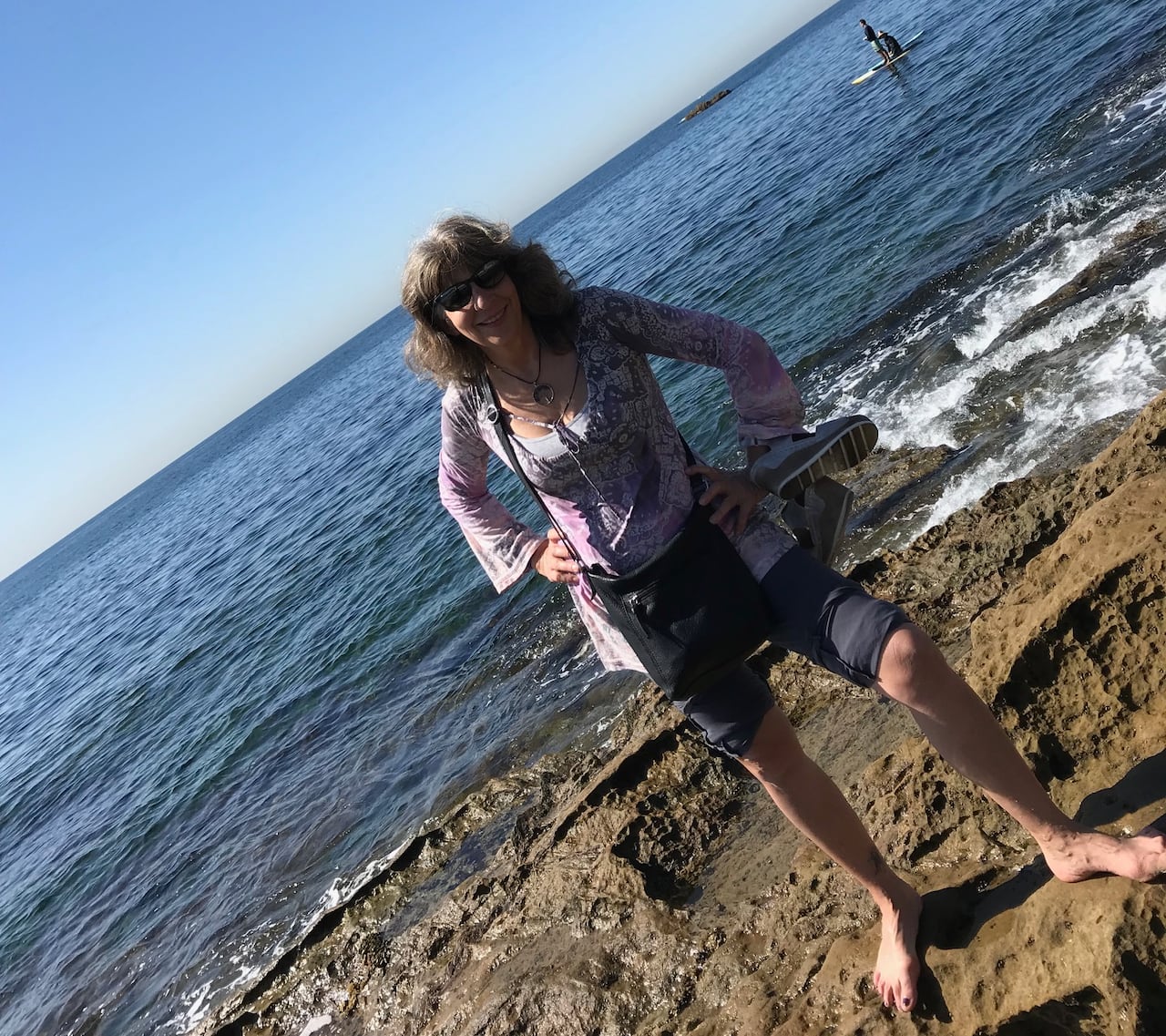 A woman wearing a long sleeved patterned blouse and denim shorts stands in the water along a beach.