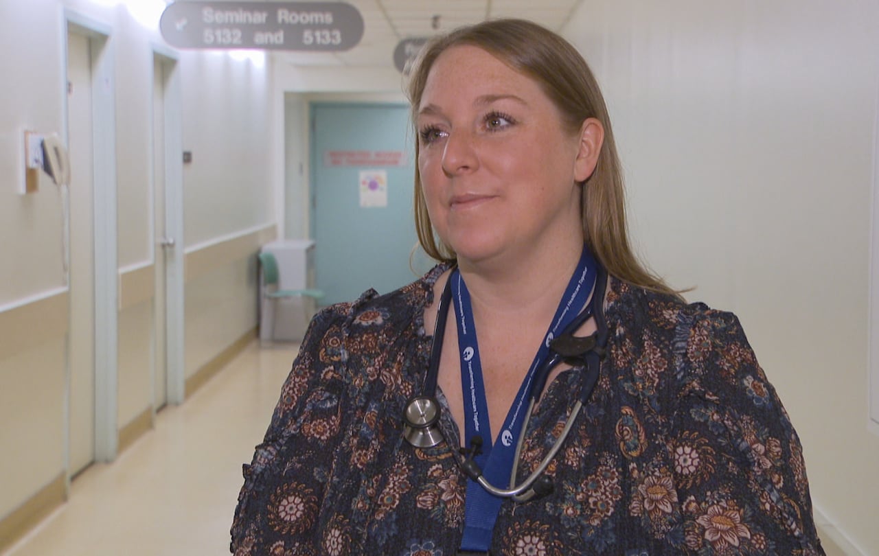 A woman with a stethoscope around her neck stands in a hallway.