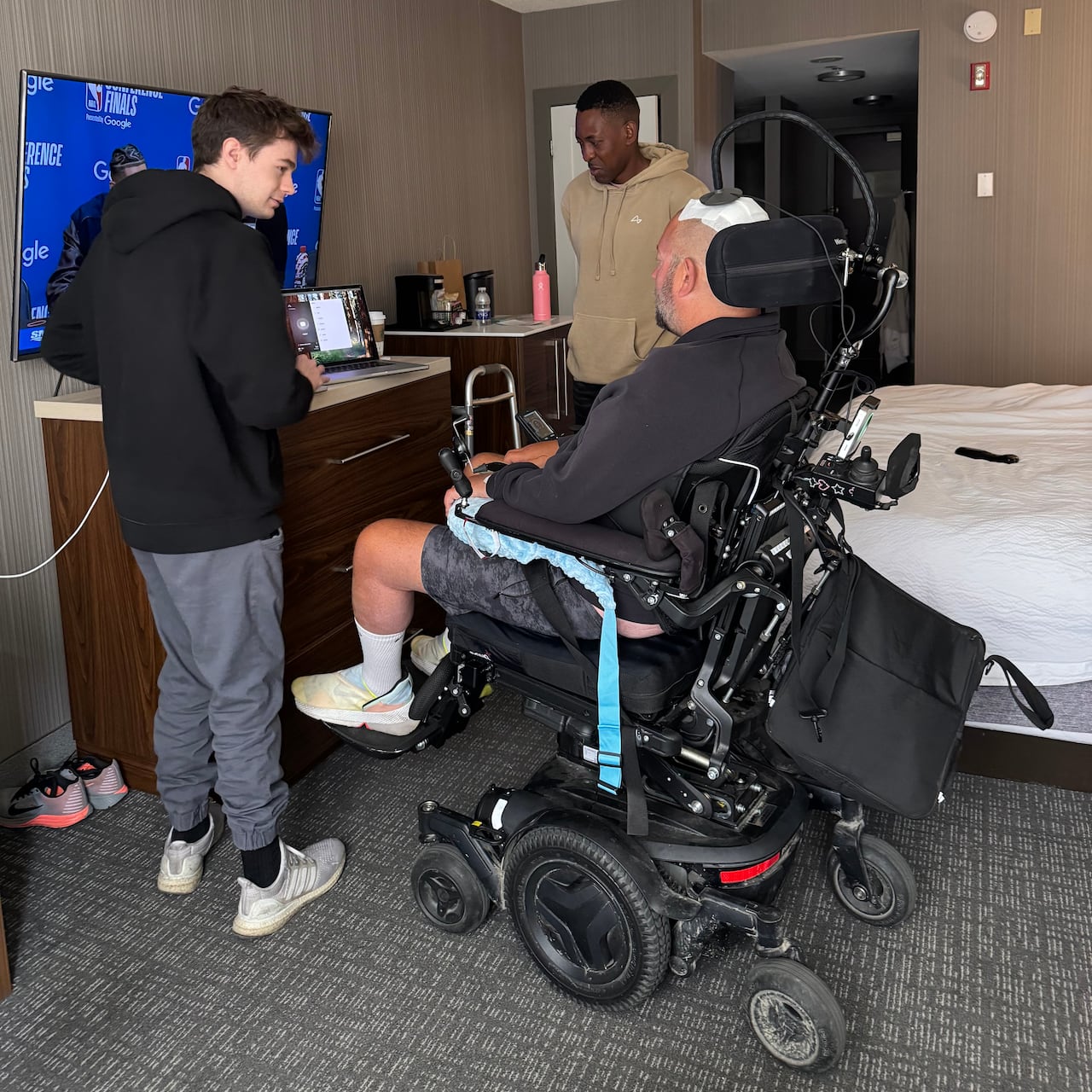 A man in a wheelchair is seen flanked by two others, as they look at a laptop positioned underneath a TV.