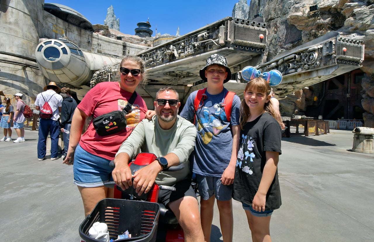 A family of four — with a man seated on a mobility scooter, a woman, a boy and a girl — smile in front of a hangar.
