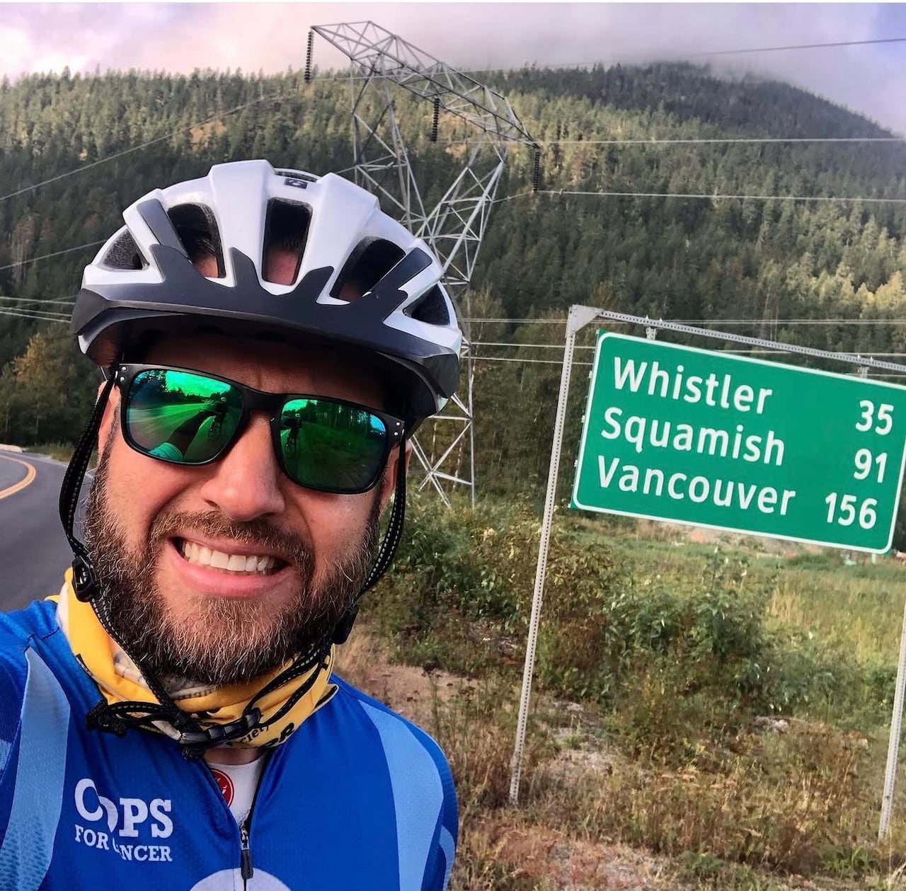 A man wearing a bicycling helmet and jersey smiles next to a road sign in the mountains.