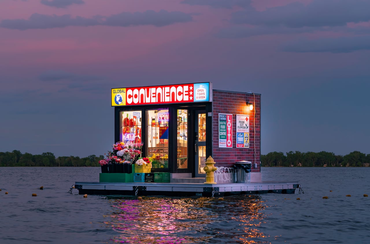 Photo of a floating art installation that replicates a city convenience store. It floats on Lake Ontario at dusk and is illuminated by shop and sign lights.