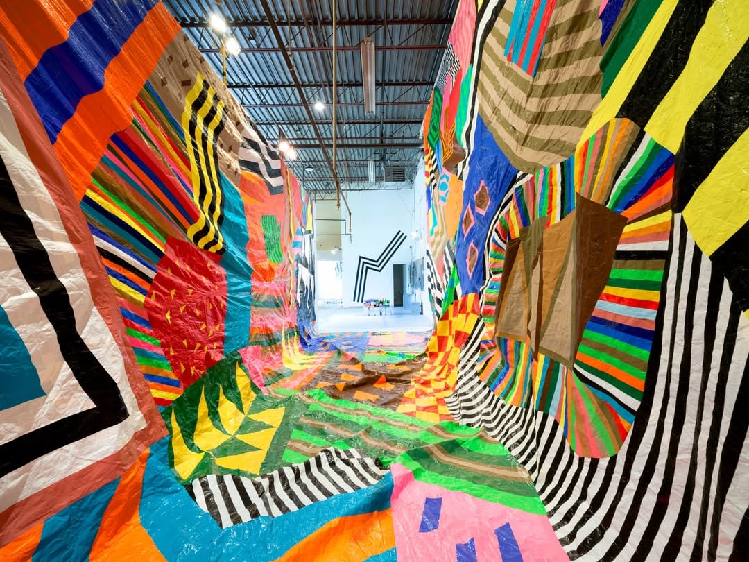 Enormous fabric artwork in a bright abstract pattern is draped from floor to ceiling in an industrial space creating a tunnel shape.