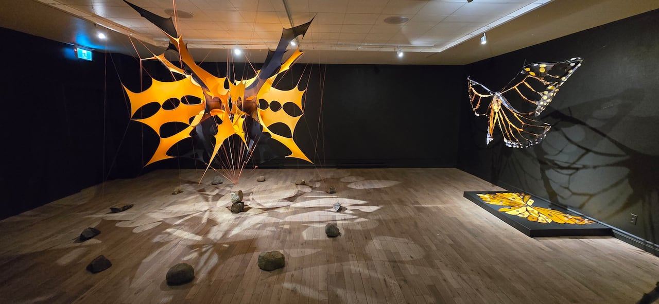 Installation view inside a dark walled gallery. Two large art installations made of fabric hang in the room. They are shaped like orange butterflies with their wings outstretched and appear to be tethered by rocks.