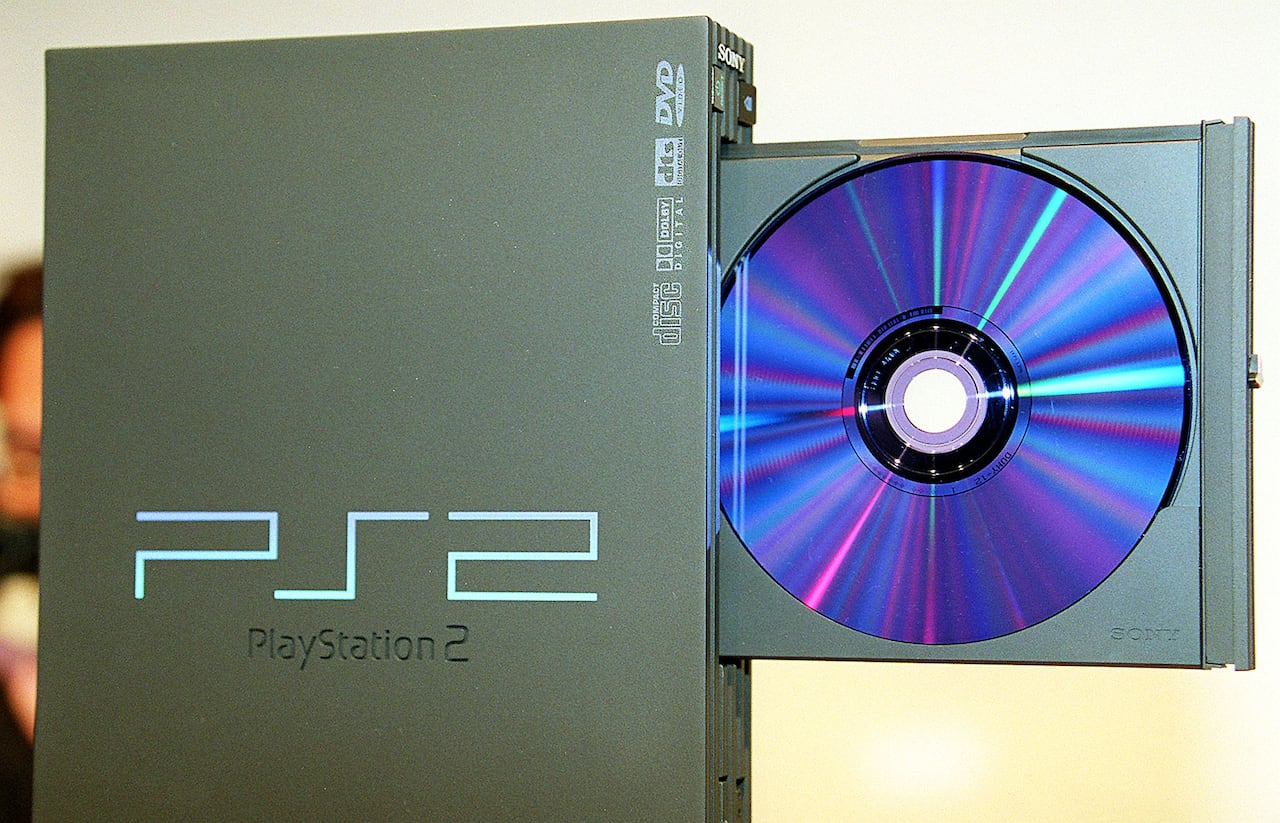 A black plastic PlayStation 2 game console is seen standing upright, with its disc tray open, showing a disc with shimmering blue colouring.