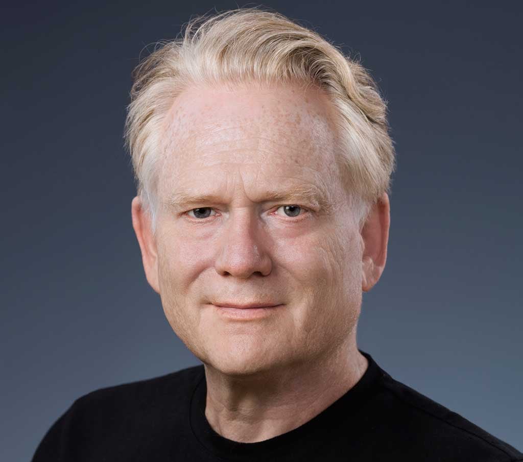 A headshot of an older man with white hair, wearing a black T-shirt.