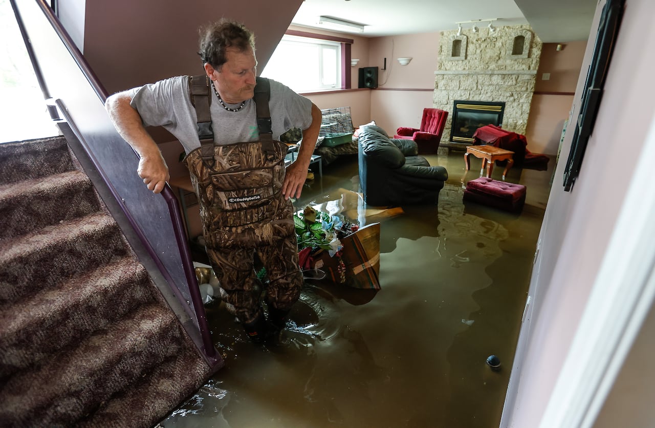 A man in overalls and a grey T-shirt stands in his basement, which is flooded.