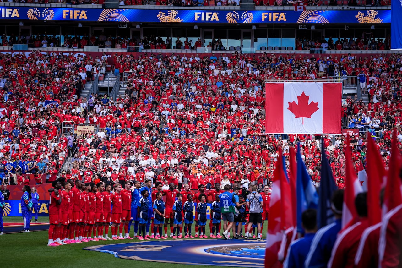 At BMO Field, Canada and Bosnia's soccer teams stand on the field ahead of a pre-game ceremony. Crowd is full and wearing red. A Canadian flag hangs in the foreground