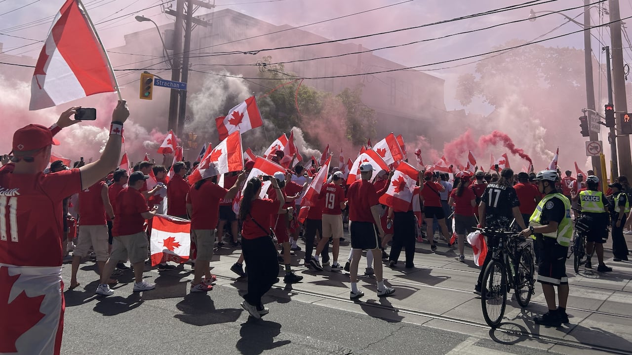 A crowd of hundreds, with thousands more off camera, march down Strachan Avenue in downtown Toronto on a sunny summer day, waving Canada flags as police block traffic