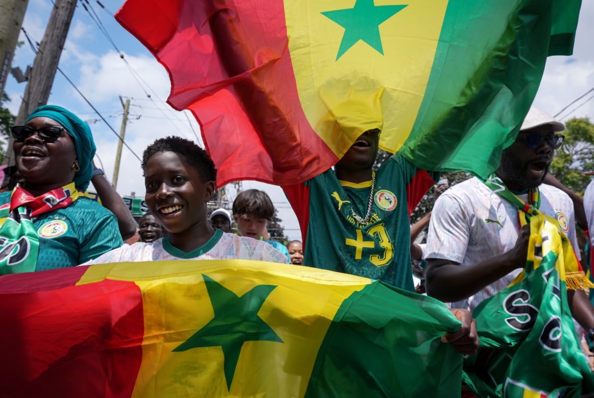 4 Senegal soccer fans in full Senegal gear wave flags at the camera as they march with a crowd through downtown Toronto on a sunny summer day.