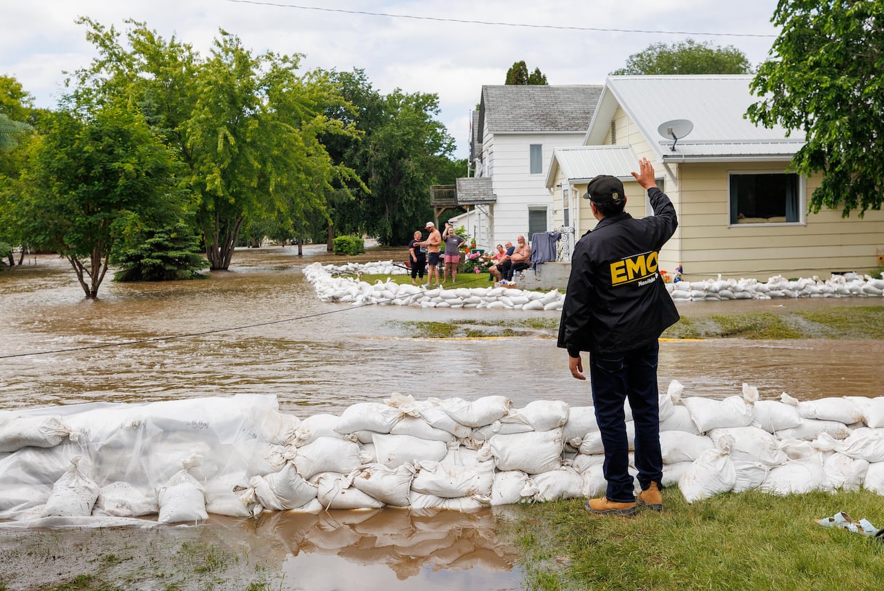 A man in a windbreaker waves across a flooded street to people sandbagging in a front yard.
