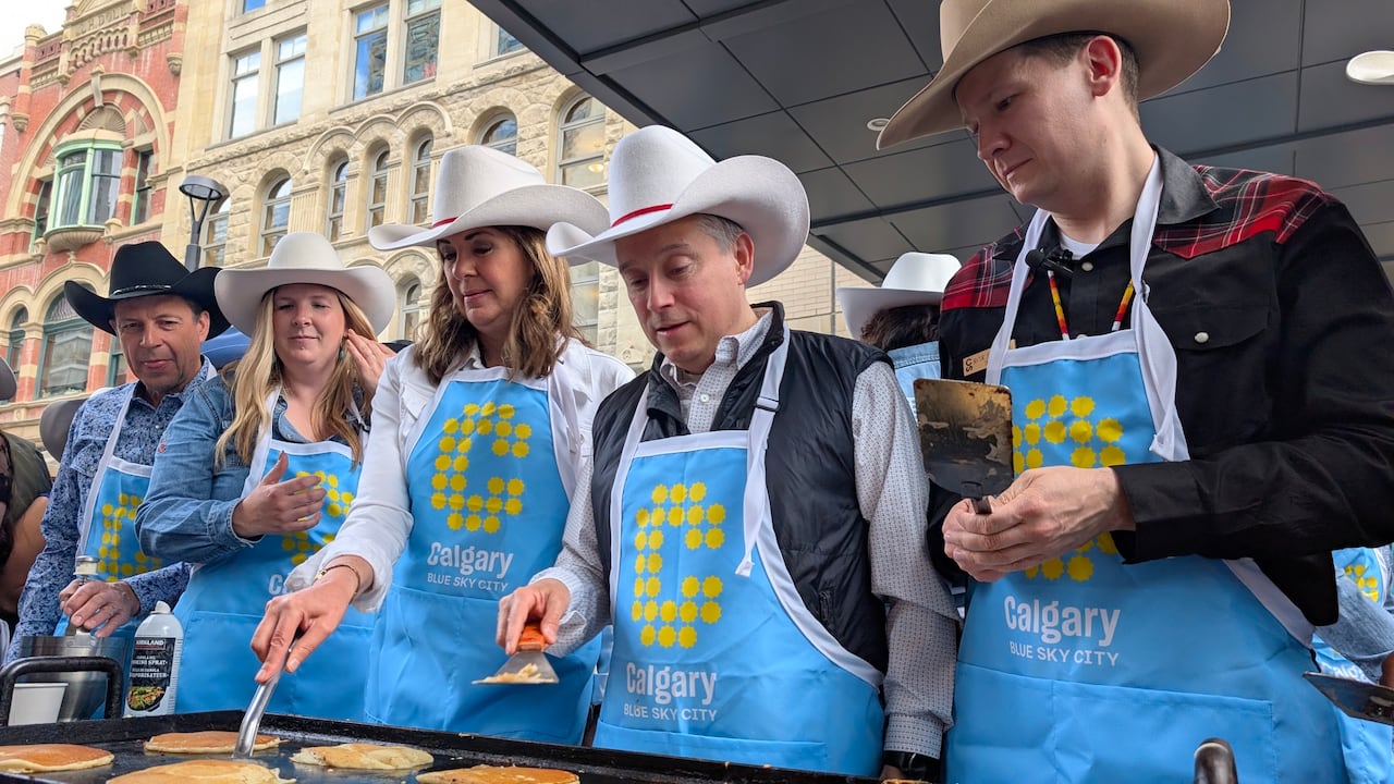 Two women and three men stand in front of pancakes on a griddle. They are wearing cowboy hats and light blue aprons with big yellow C's on them.