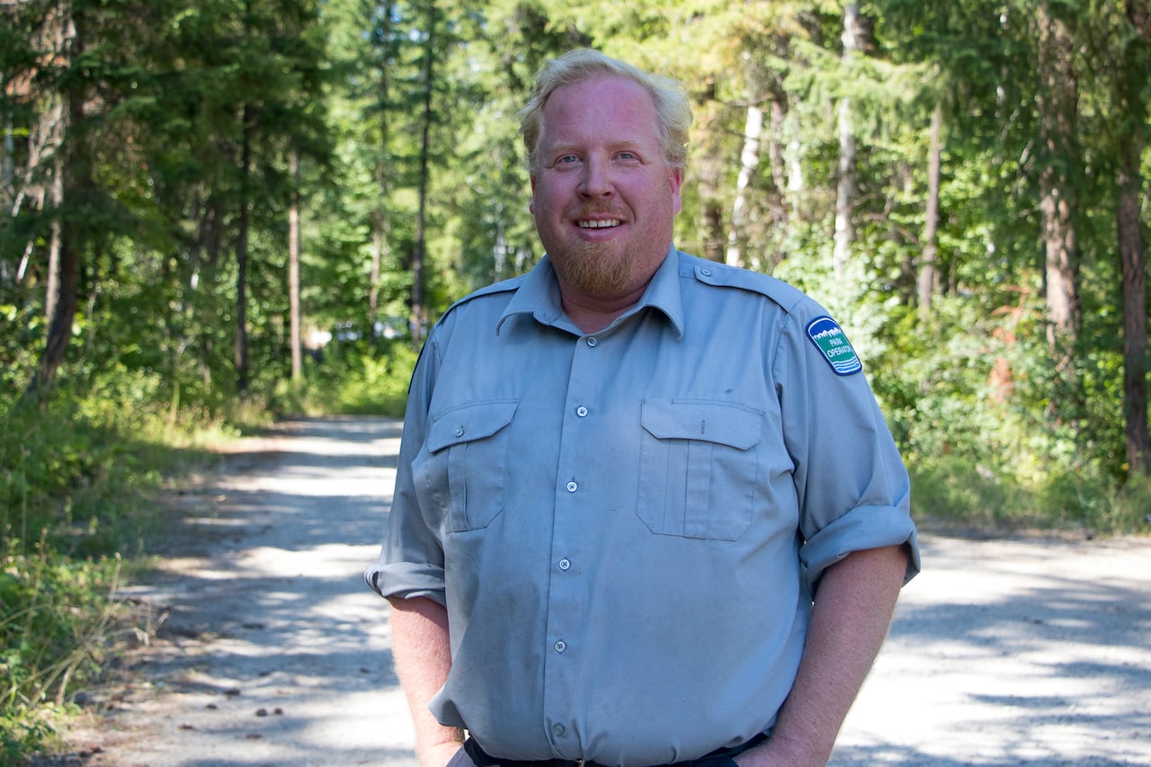 A man with a light red beard and blond hair in a blue open-collared shirt smiles at the camera framed by the forest behind him.