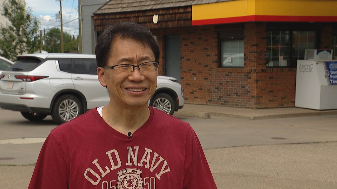 A man of East Asian descent stands in front of a convenience store wearing a red t-shirt.