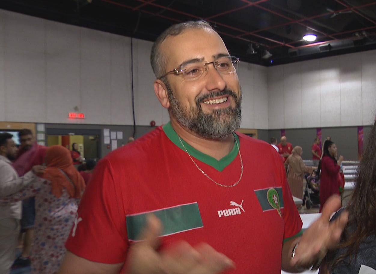 A man in a red and green jersey smiles and holds out his hands.