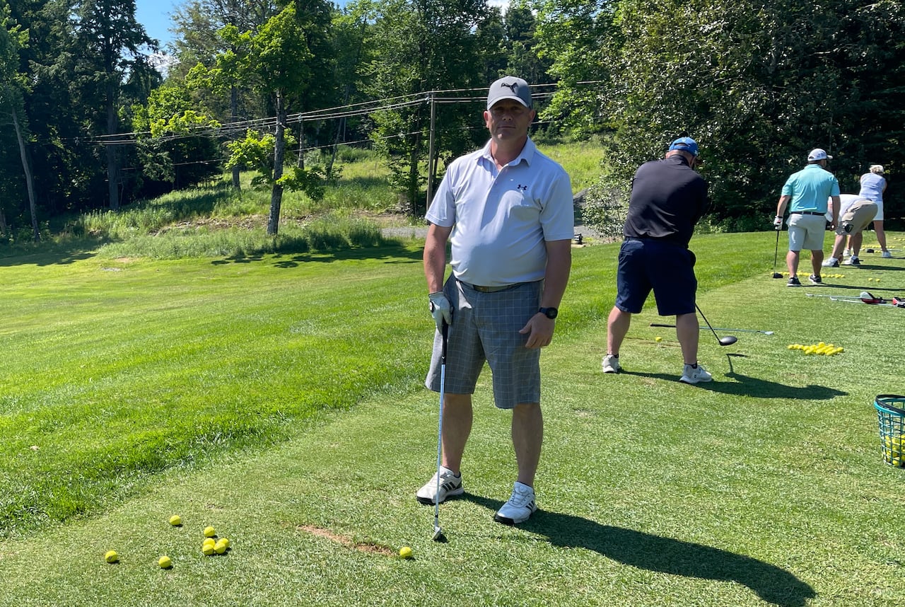 A man in a grey shirt, shorts and hat standing on a driving range with a golf club.
