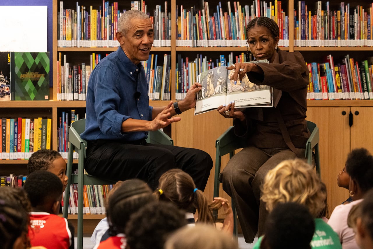 Two adults sitting on chairs read to children seated on the ground.