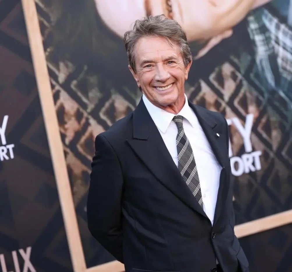 Martin Short wearing a suit and tie and smiling broadly in front of a poster for his Netflix documentary.