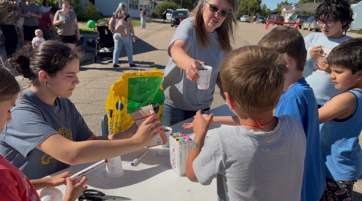 Two women help five kids plant seeds in styrofoam cups at a small town street party.