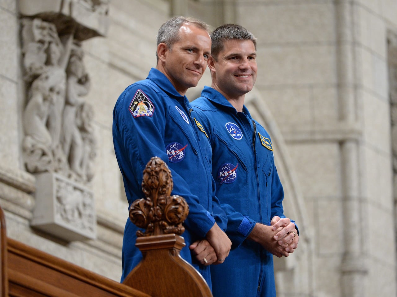 Two men stand in blue astronaut uniforms before a stone wall.