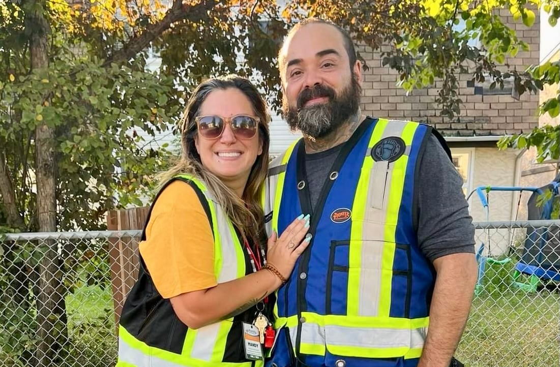 A woman and a man, both wearing reflective vests, stand on a city sidewalk.
