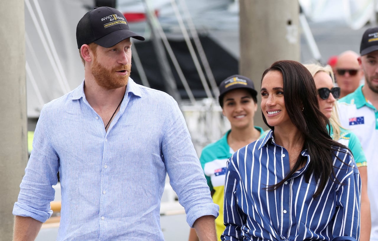 A man in a blue shirt and black baseball cap walks with a woman in a striped blue shirt on a dock, followed by a small crowd.