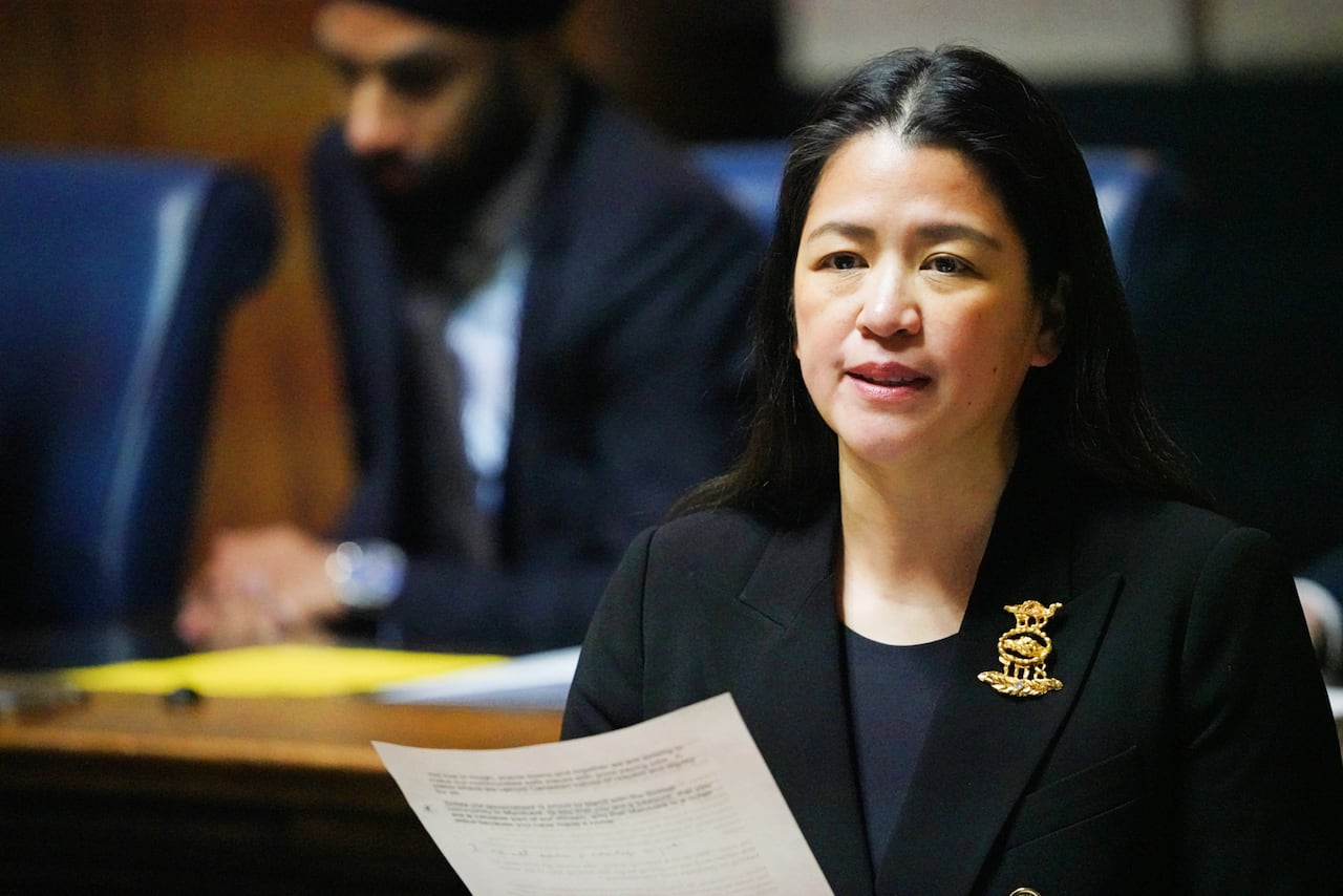 A woman with long dark hair and a black blazer speaks while holding up a paper during a government legislative session.