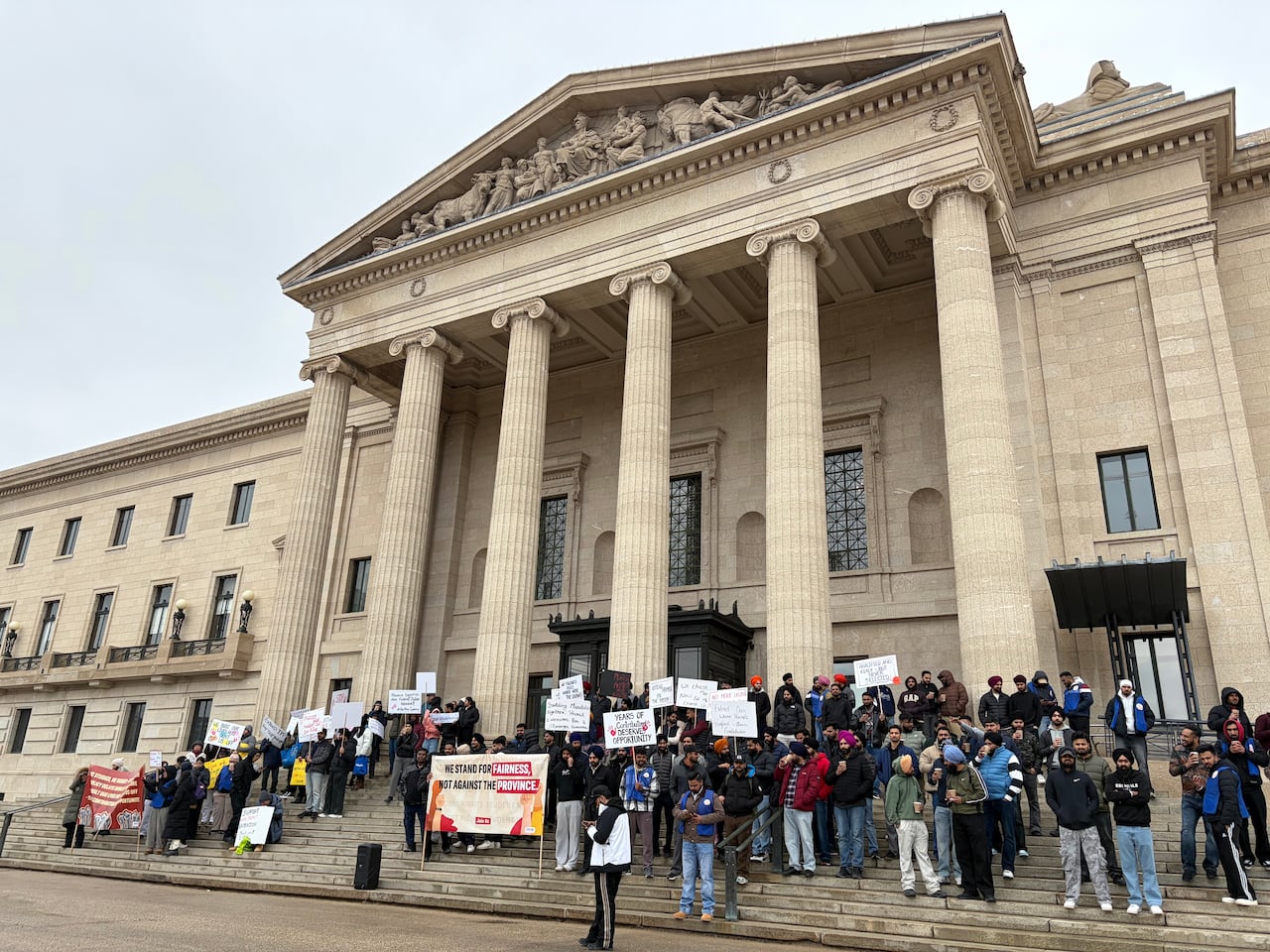 About 130 people stand on the steps of a government building holding signs during a protest.