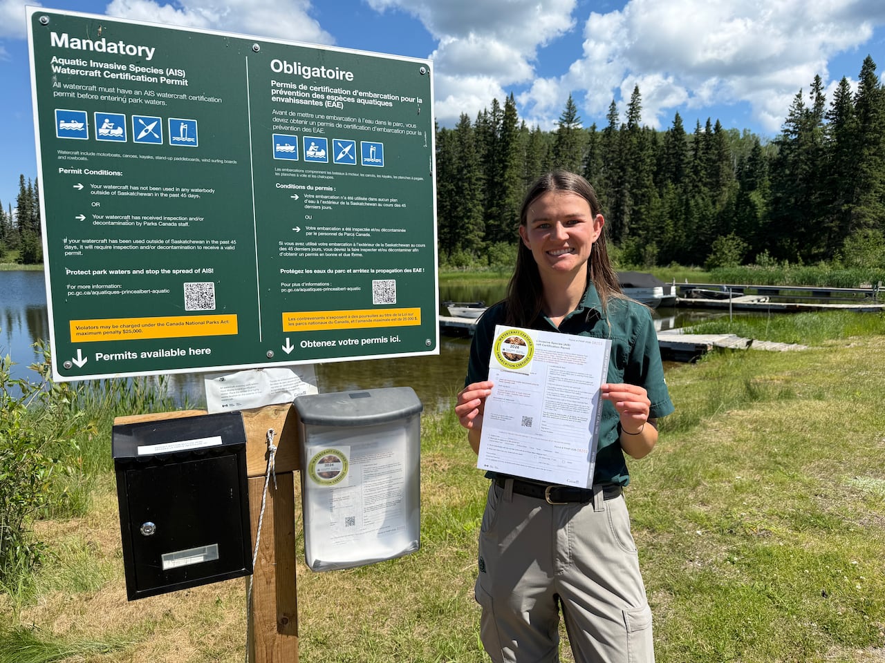 A young woman with brown hair holds a permit and sticker while standing beside a sign on the shore of a lake.