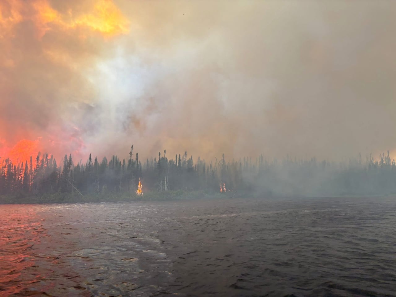 A red and orange sky is filled with smoke over a line of trees and the water of a lake.