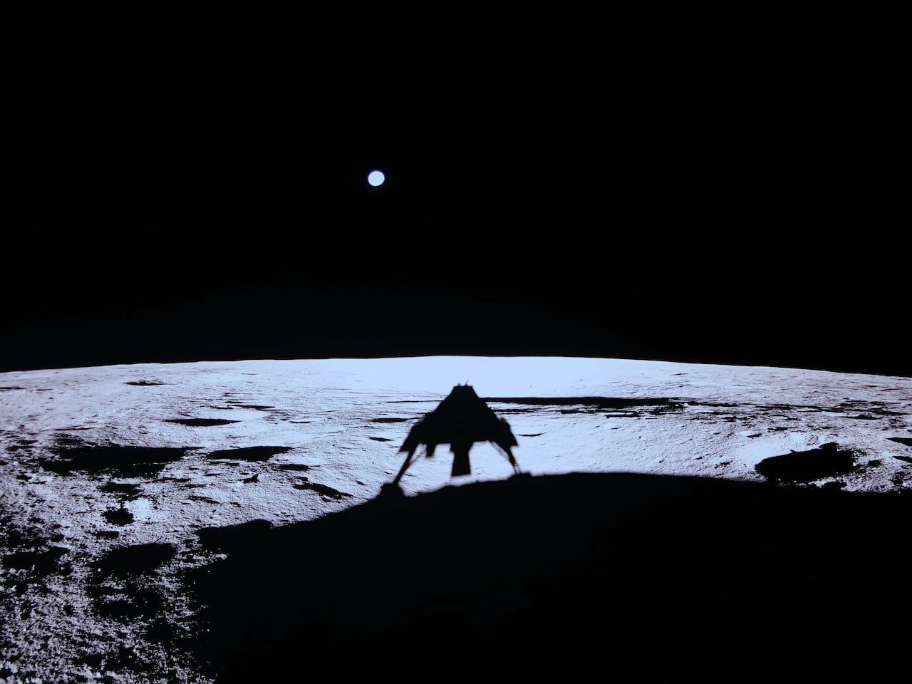 The shadow of a lunar lander is seen on the surface of the moon with Earth in the distance.
