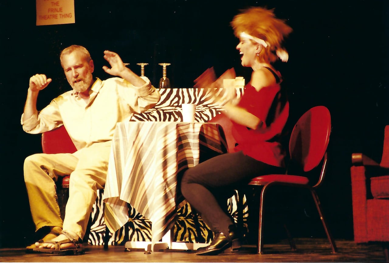 A man sitting at a table raises his arms in surprise while a woman in a red shirt laughs and moves her arms wildly. Both are sitting at a table on a stage.