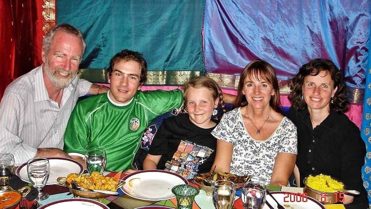 A man, younger man, teenage girl and two women sit at a dinner table surrounded by brightly coloured blankets hanging on the walls.