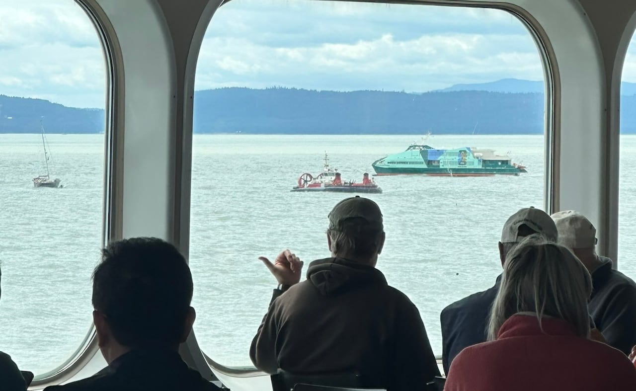A Coast Guard hovercraft, a Hullo foot passenger ferry and a sailboat seen through the window of a B.C. ferry with passengers watching in the foreground.