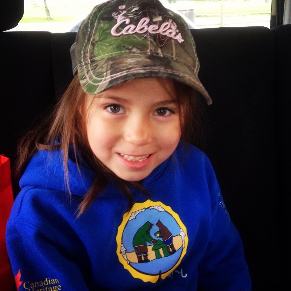 A young girl in a camoflauge cap and a blue sweater smiles at the camera.