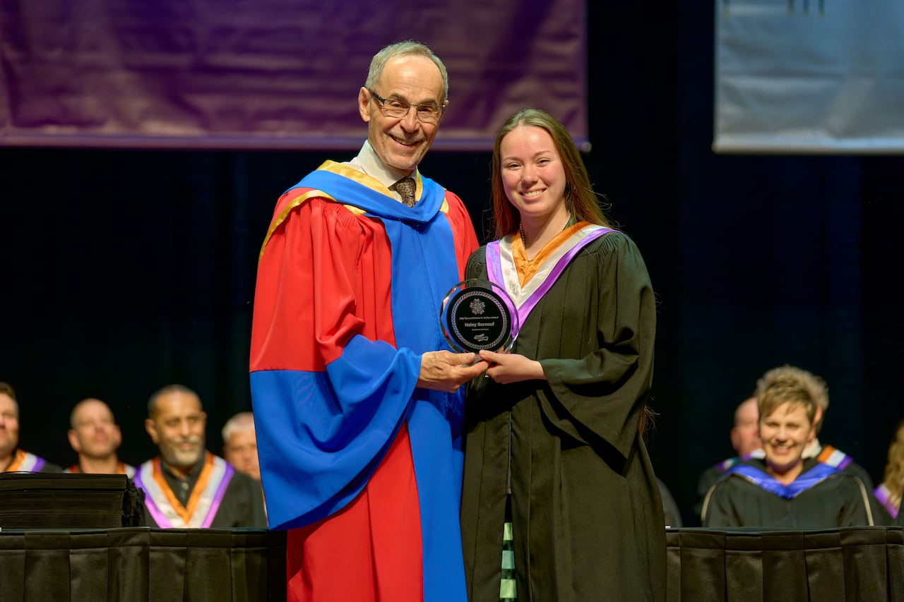 A man in blue and red robes presents a glass plaque to a woman in a black graduation robe.