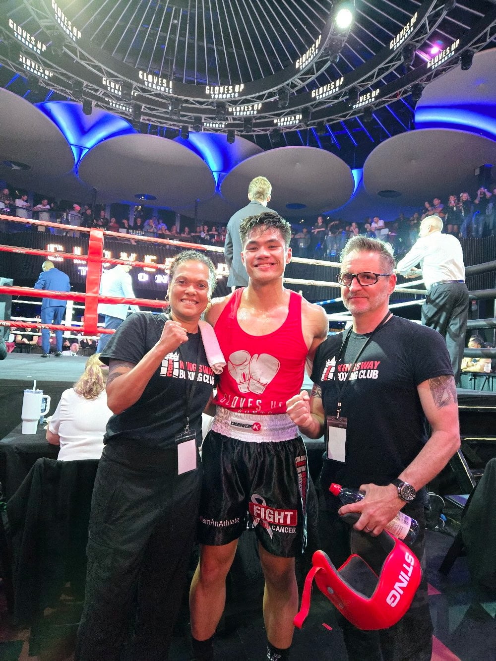 Two coaches stand on either side of a boxer, in front of a boxing ring.